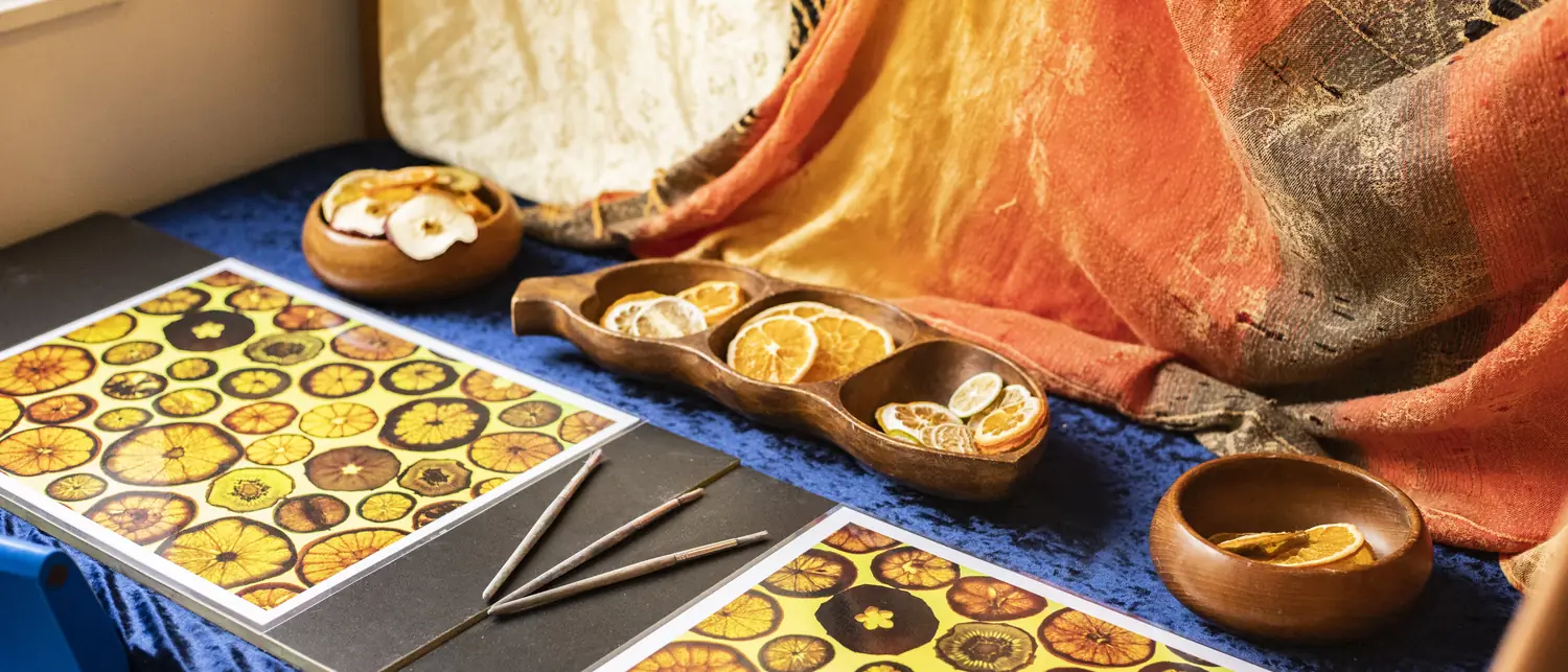 A table draped in blue and orange cloth, with wooden bowls of dried orange slices, fine paintbrushes and printed sheets of fruit slices.