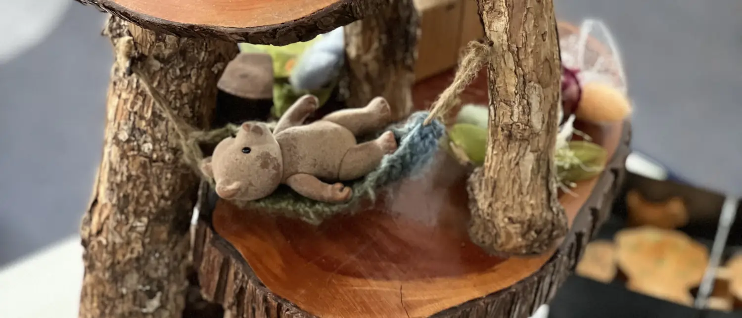 A handmade wooden tree house of log slices, with a small felt bear lying in a knitted hammock.
