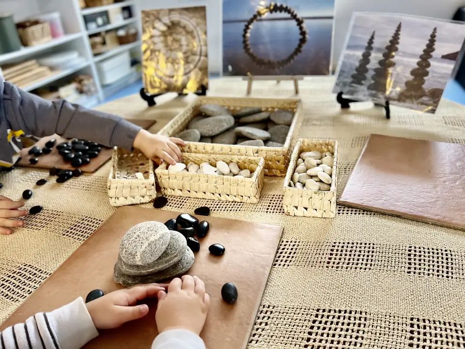 Children's hands sorting black stones and pebbles from woven baskets on a lace-covered table, with photographs of stone patterns propped behind.