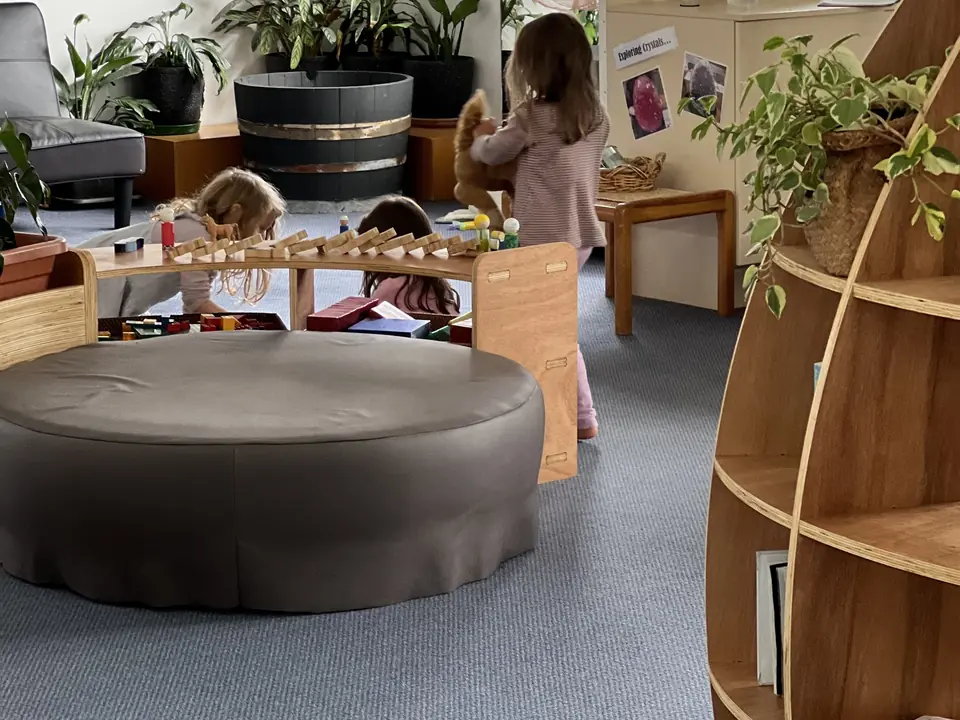 A round grey ottoman beside a low shelf of blocks, where three children play with their backs to the camera, and a tall curved bookcase in front.