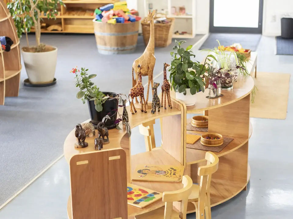 A curved wooden shelf with carved giraffes and elephants, plants, bowls and wooden puzzles, and two small chairs.