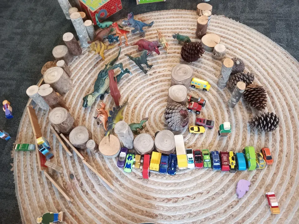 A spiral of loose parts on a round woven mat: pine cones, wooden pegs, toy cars and dinosaurs.