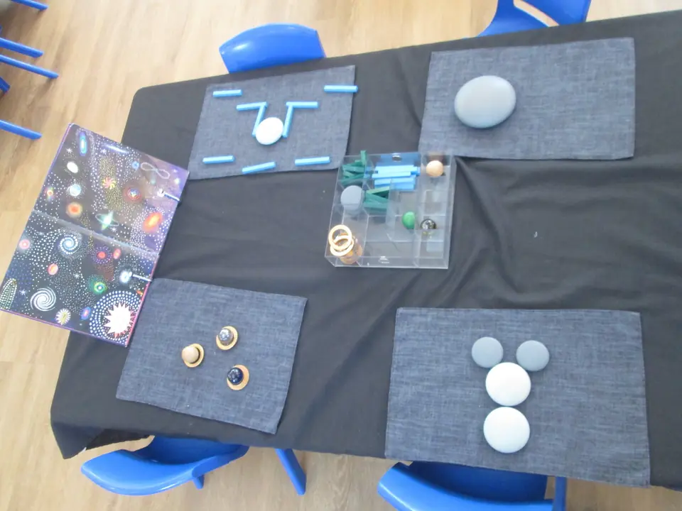 From above: a black table set with mats, stones, rings and a clear box of space pieces, with blue chairs around it.