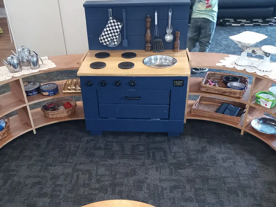 A blue play kitchen with utensils hanging from its backboard, set in a half circle of curved wooden shelves holding teapots, tins, baskets and pans.