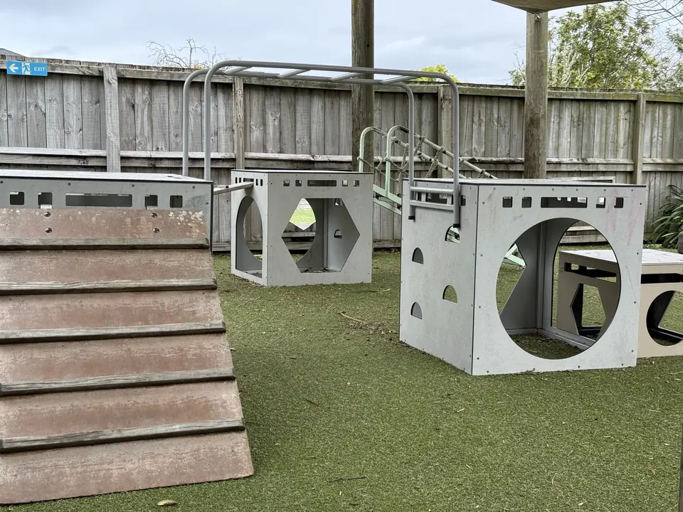 White play cubes with cut-out shapes, a wooden ramp and a climbing frame on turf, inside a timber fence.