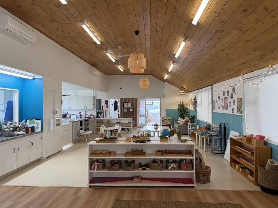 A long room under a timber ceiling, with a low white shelf of materials in front, a kitchen bench along one side and wooden shelves beyond.
