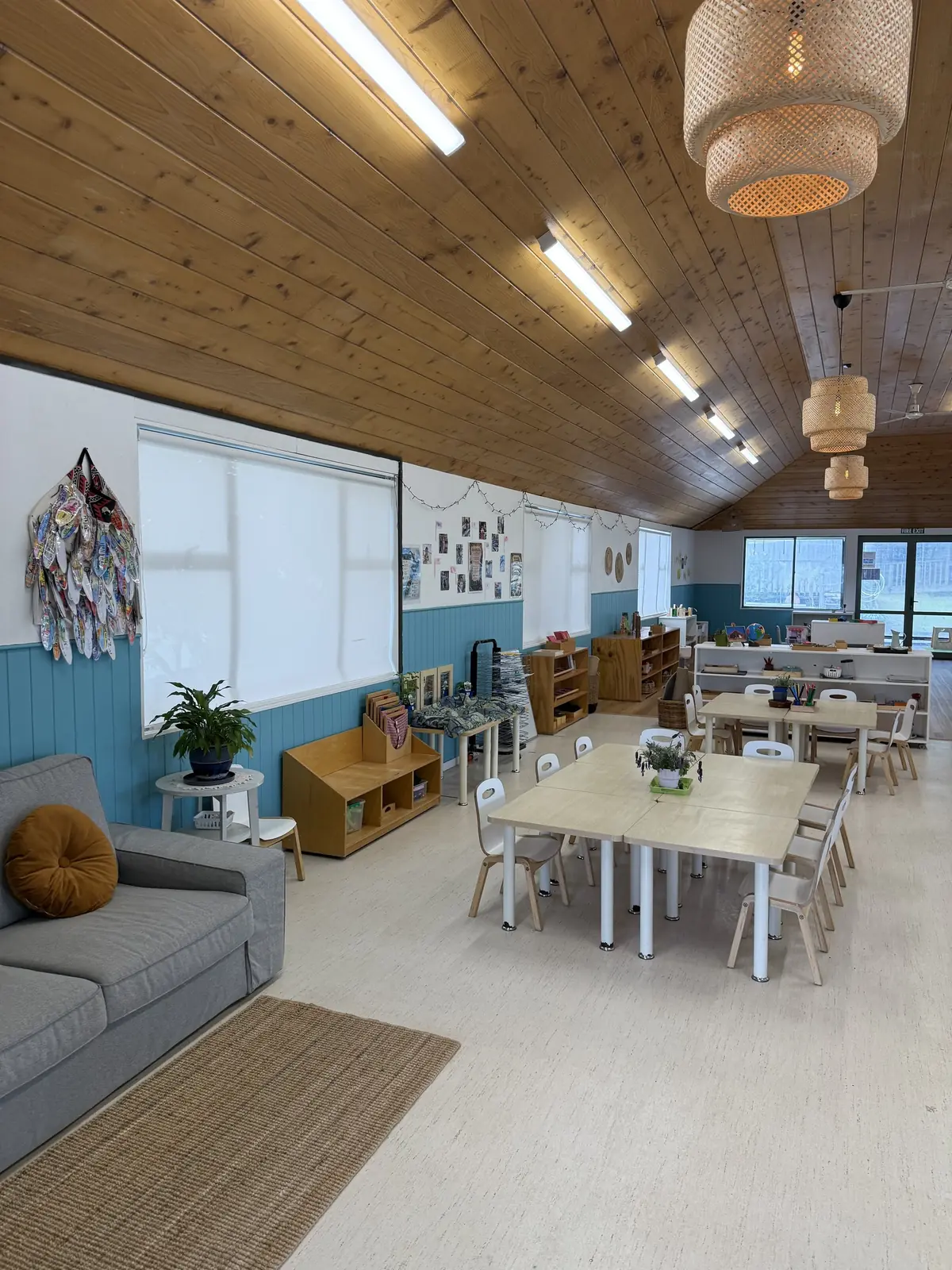 Small tables set with plants in a room with teal panelling, a grey sofa with an orange cushion, and woven pendant lights overhead.