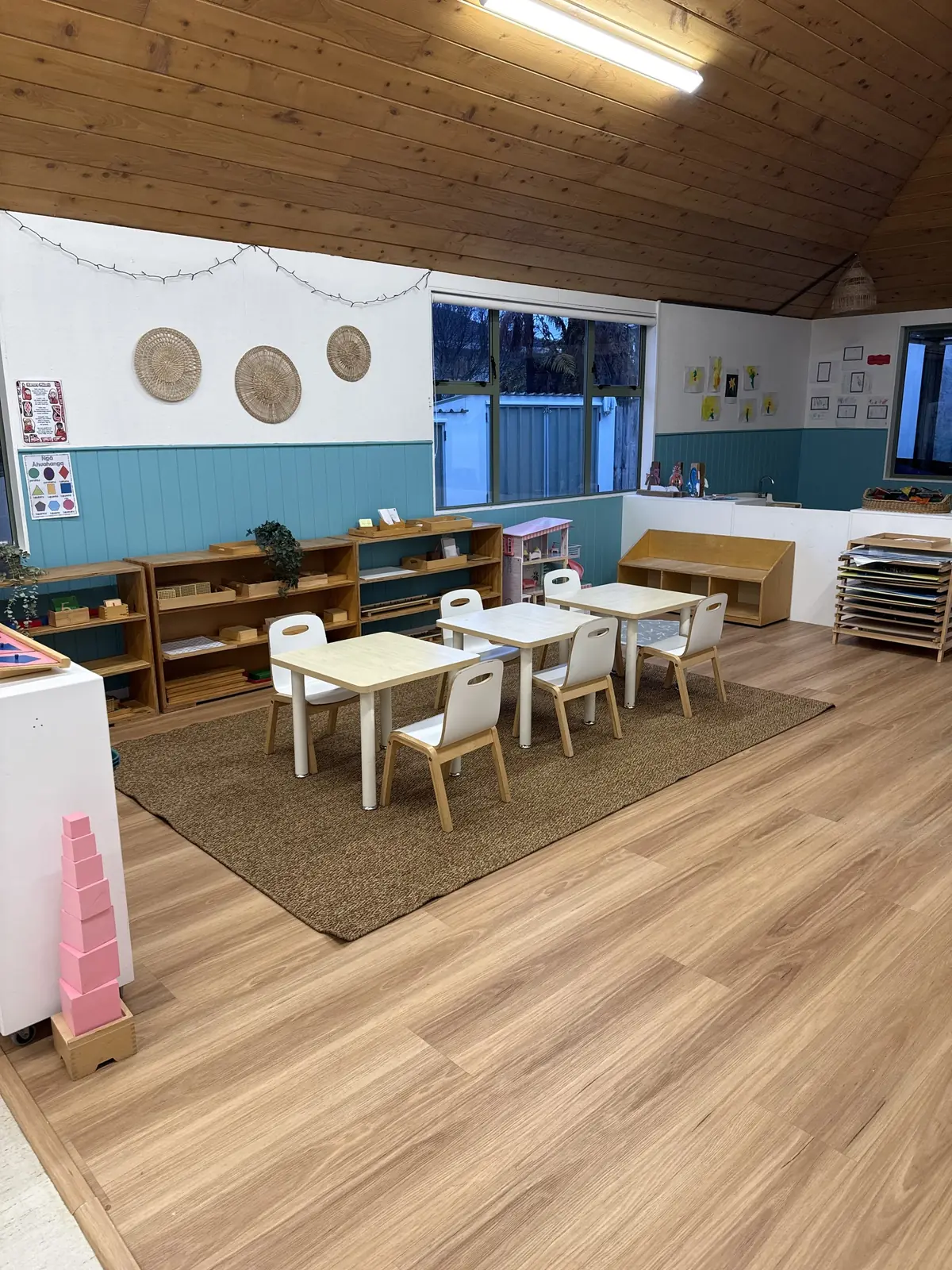Small white tables and chairs on a jute rug in a room with a timber floor, wooden shelves of materials along teal panelling and woven baskets on the wall.