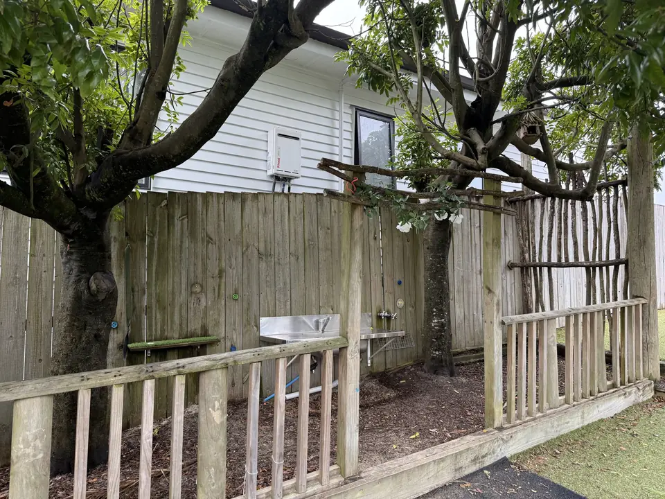 A fenced garden corner under two trees, with a low wooden railing in front and an outdoor sink on the fence.