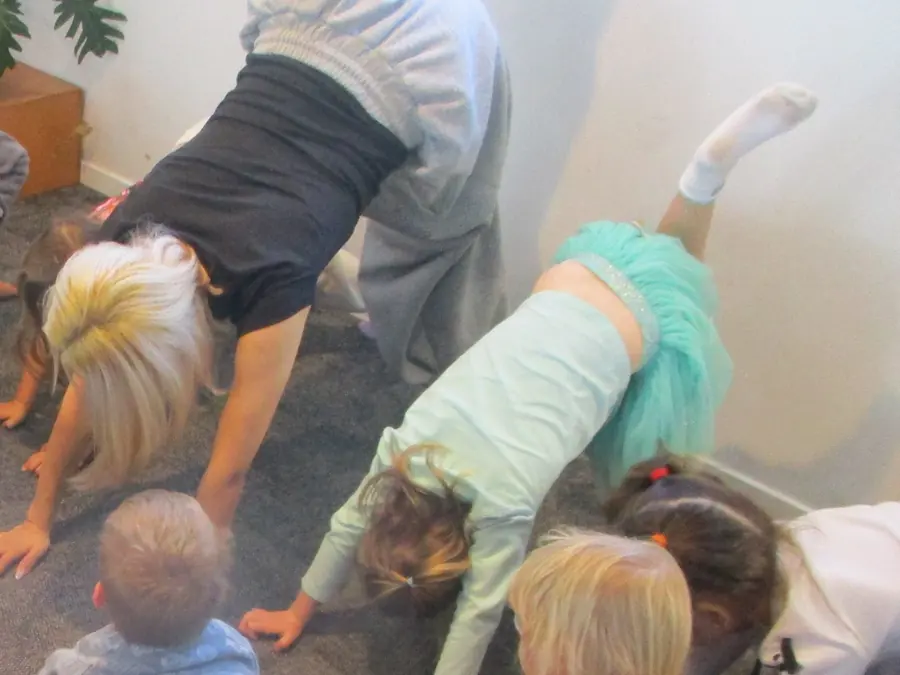 A movement session on the carpet: a teacher and a girl in a green tutu stretch with their heads down, other children around them.
