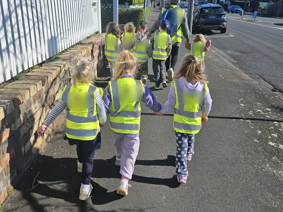 Children in hi-vis vests walk along a footpath holding hands, a teacher ahead of them.