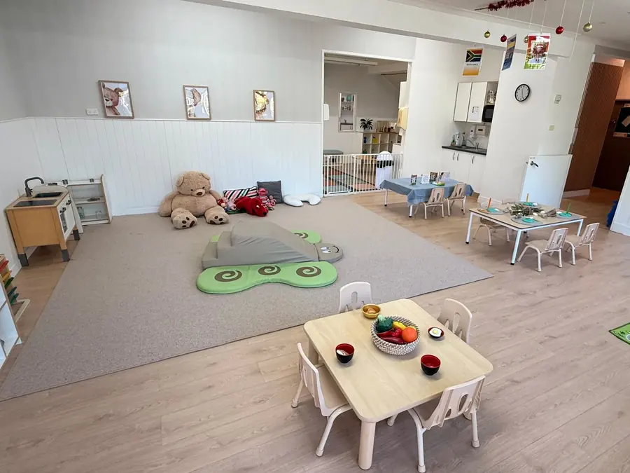The babies' room at Anzac Street, Takapuna, wide: a large teddy, soft-play shapes, small tables and a play kitchen.