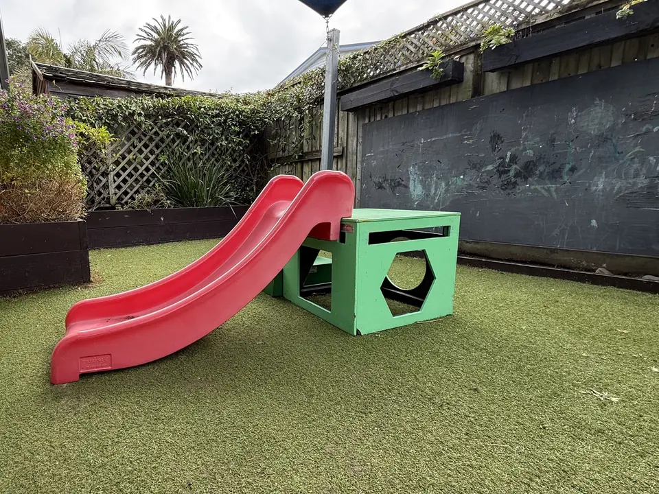 A red slide and a green play cube on turf, beside planter boxes and a chalkboard wall along the fence.