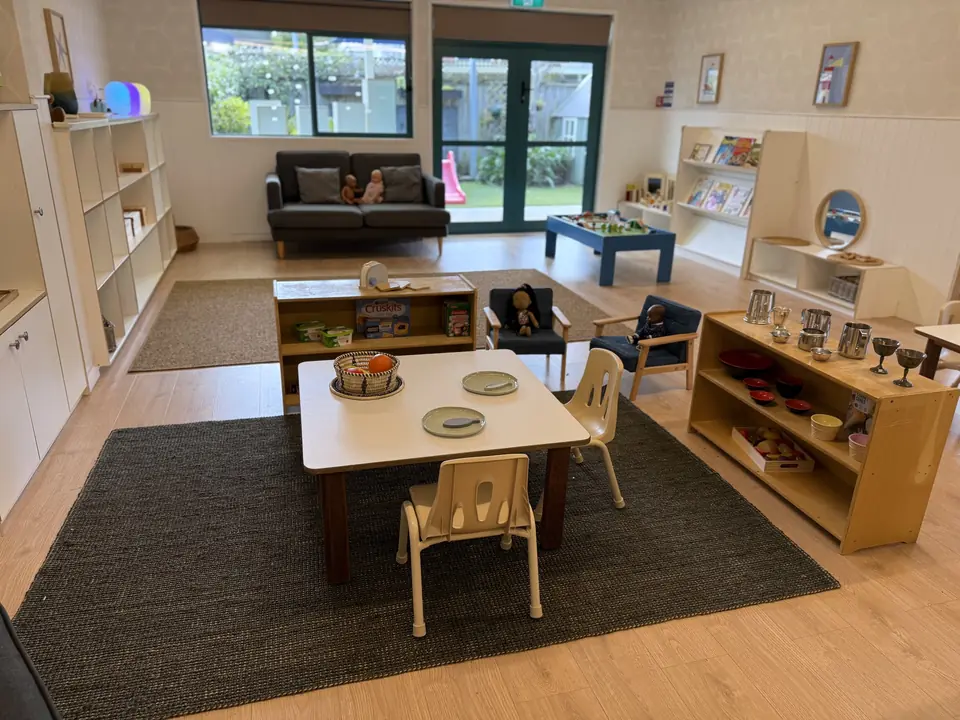 A small table set with plates on a dark rug, blue armchairs with dolls, low wooden shelves and a sofa by glass doors.