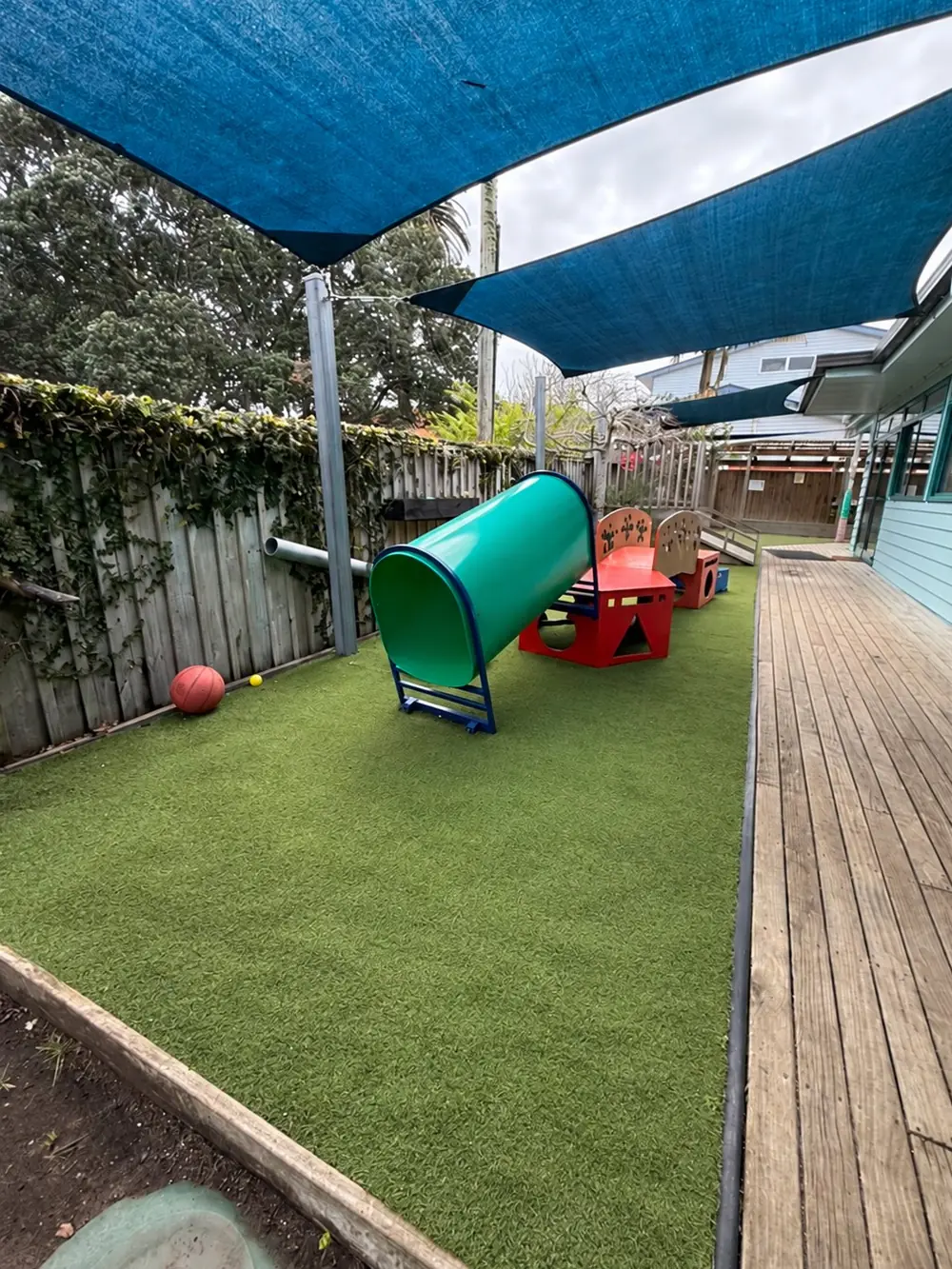 A strip of turf beside a timber deck, with a green tunnel, red play shapes and a ball, under blue shade sails.