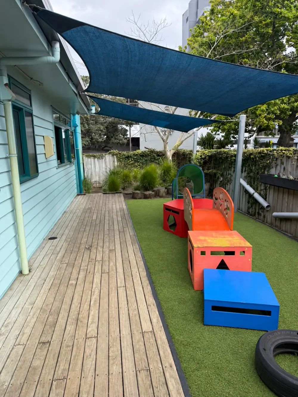 A timber deck along the building beside a strip of turf, with orange, red and blue play blocks and a tyre under blue shade sails.