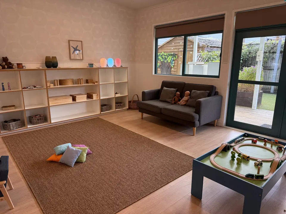 A room with a grey sofa and dolls under the window, a wooden train table, low shelves of materials and scatter cushions on a jute rug.