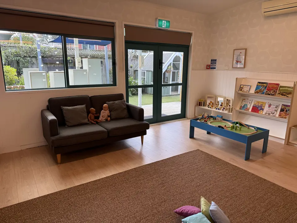 A grey sofa with dolls and cushions, a blue train table and a rack of picture books, with glass doors and windows to the yard.