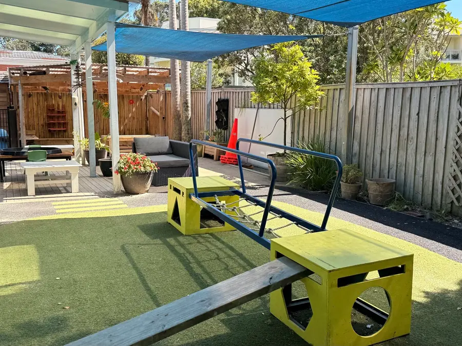 The main yard at Anzac Street, Takapuna: a yellow balance beam and platforms on turf under blue shade sails.